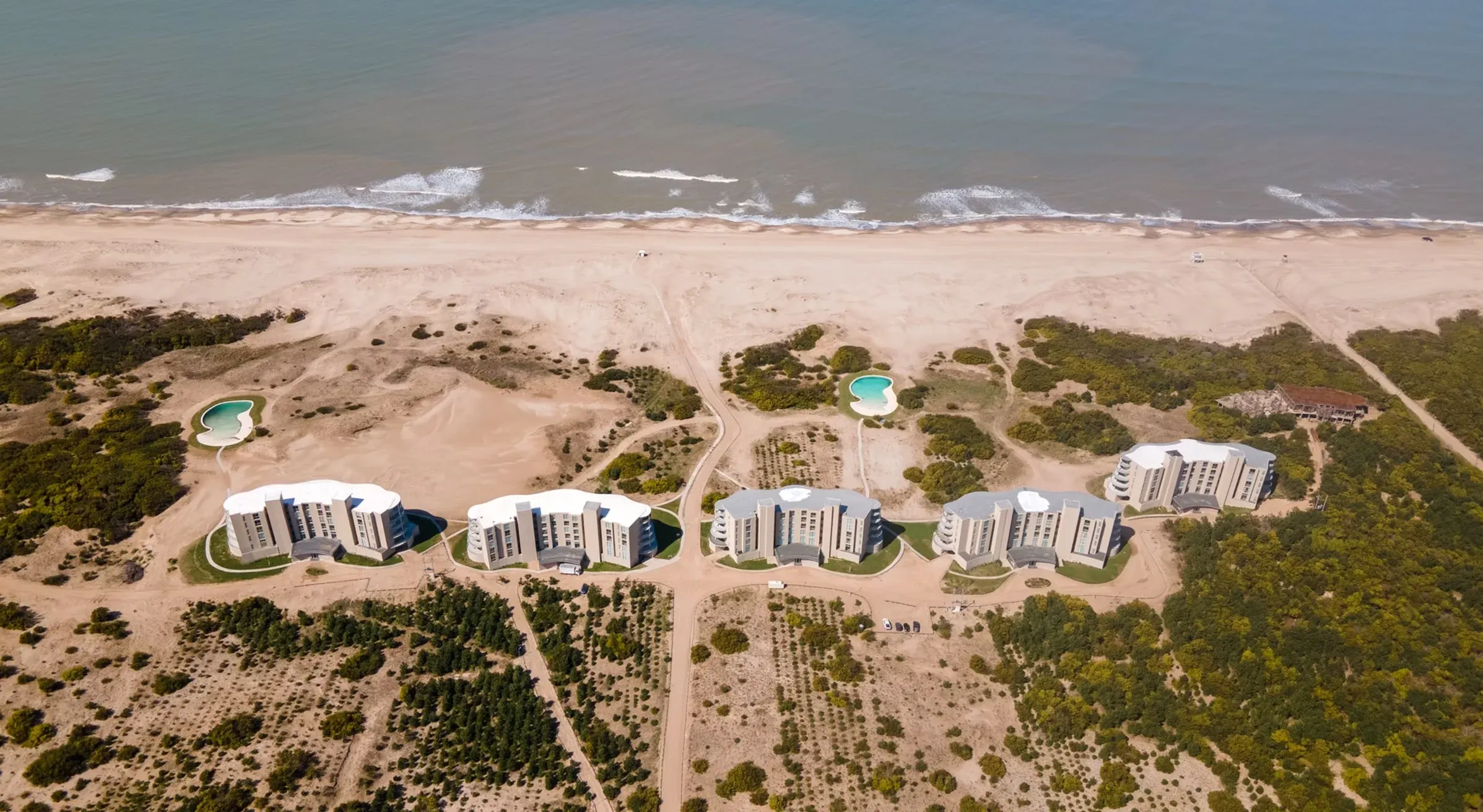 Vista aérea real de cinco edificios de NorthBeach entre dunas y mar, mostrando la escala del desarrollo
